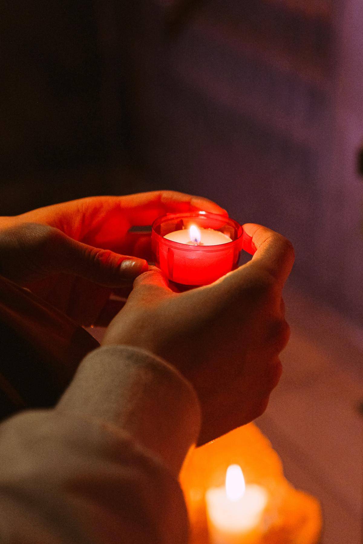 Quelle: pexels.com / Anna Shvets, https://www.pexels.com/de-de/foto/licht-rot-hand-dunkel-3943924/ Kerze in Kirche wird in Händen gehalten